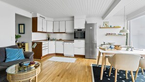Een moderne keuken met witte kasten, een roestvrijstalen koelkast en een inbouwoven. Er is een eethoek met een tafel en stoelen, een bank en een salontafel met fruit en glazen.