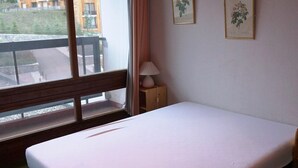 Una habitación con una cama individual, una ventana con vistas a edificios, una lámpara en una mesita de noche y dos cuadros en la pared.