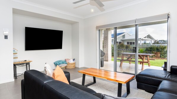 A modern living room with a flat-screen TV, black leather sofas, a wooden coffee table, and a sliding glass door leading to an outdoor patio with a picnic table. - Surf and Dunes- Yamba (Yamba)