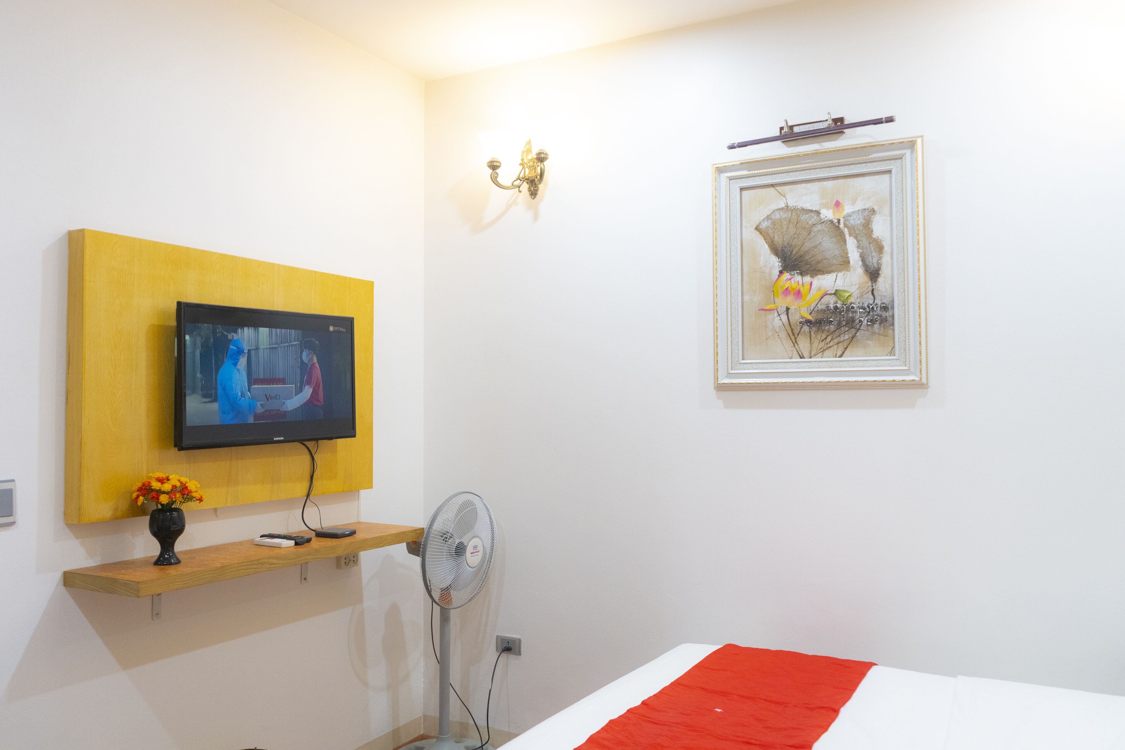 Una habitación de hotel con una televisión montada en una pared amarilla, una obra de arte enmarcada sobre la cama y una mesita de noche con un jarrón de flores.