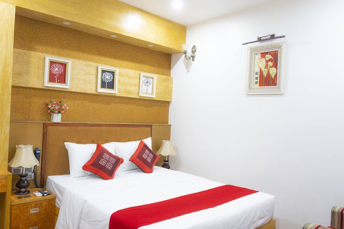 A hotel room with a wooden headboard, a bed with white bedding and red accents, a nightstand with a lamp, a small table with a vase of flowers, and framed pictures on the wall.