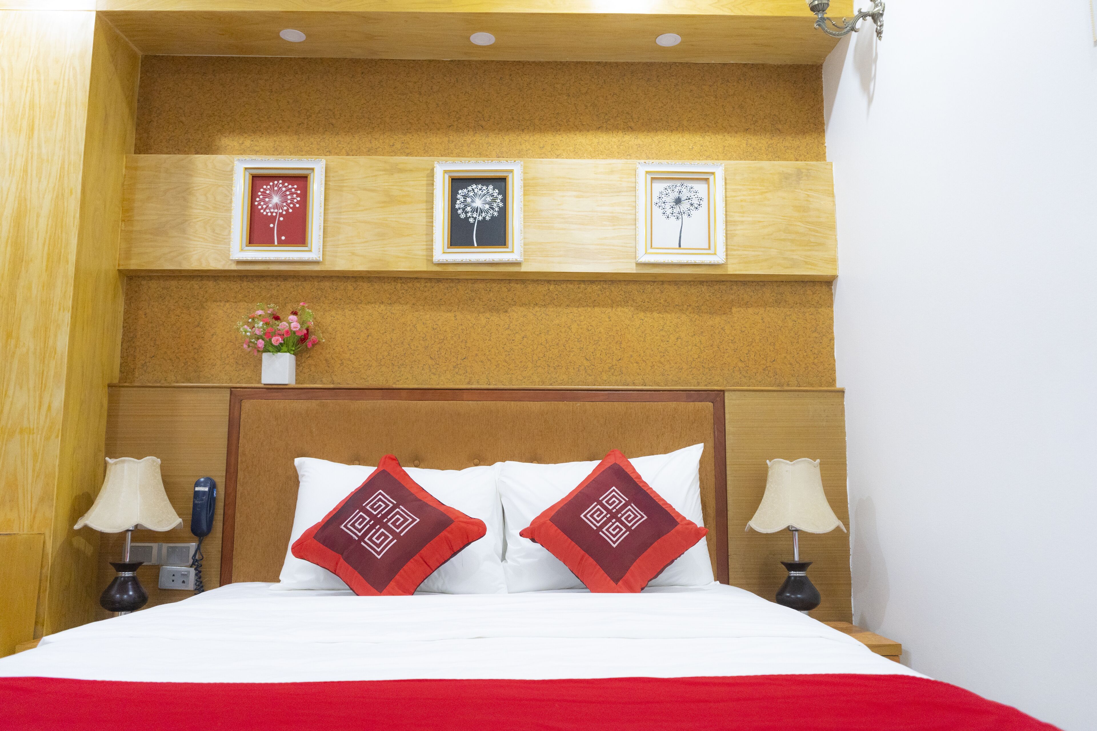 A hotel room with a wooden headboard, a bed with white and red bedding, two bedside lamps, a phone on the nightstand, and three framed pictures on the wall.