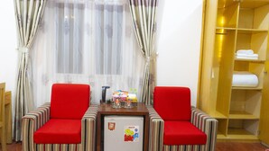 Habitación de hotel con cama, dos sillones a rayas, un refrigerador pequeño, un armario de madera y una ventana con cortinas translúcidas.