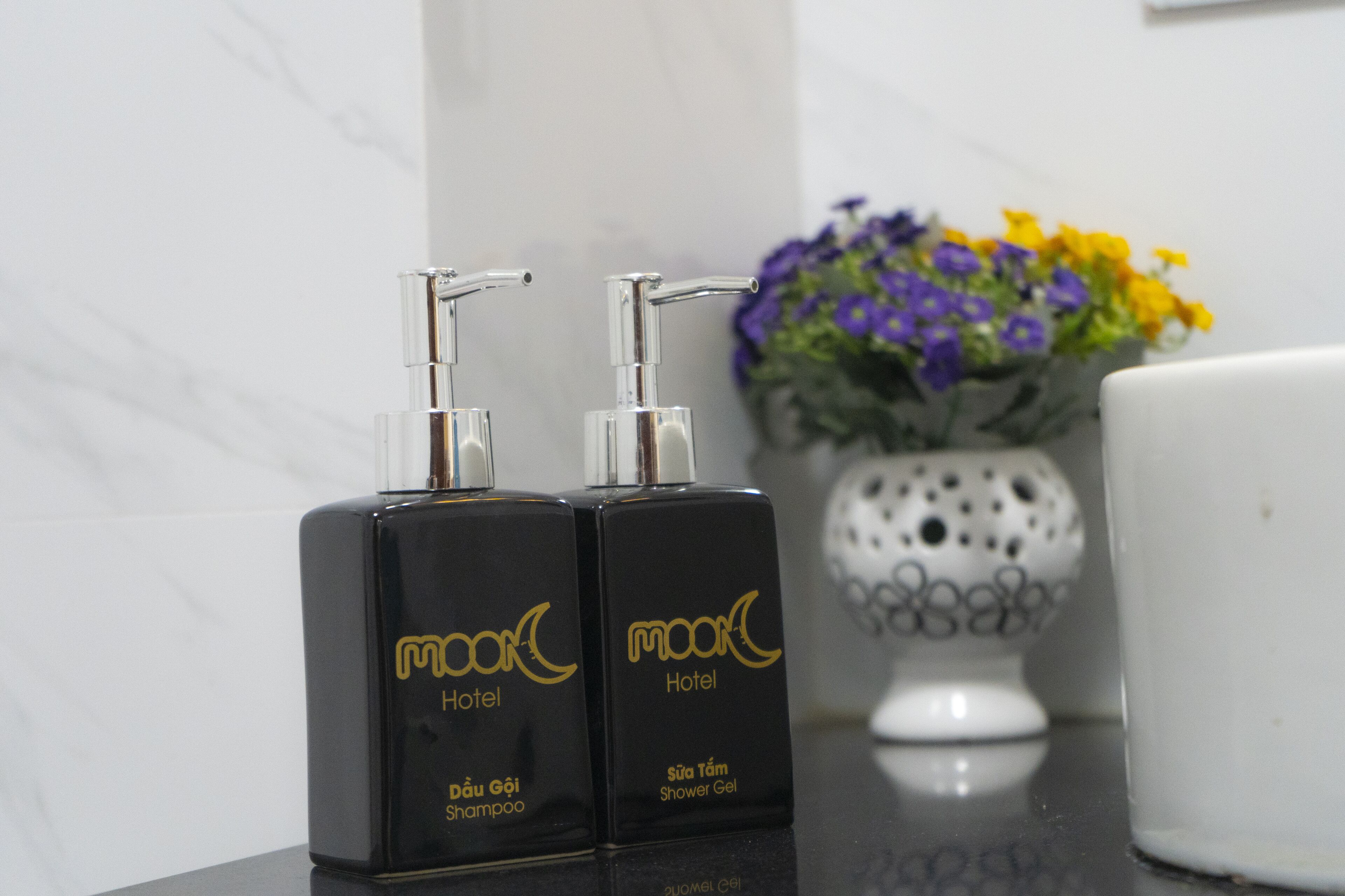 Two black bottles of MooC Hotel shampoo and shower gel on a reflective surface.
