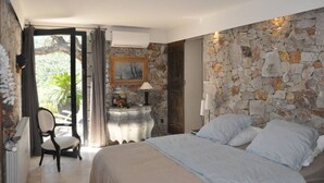 A bedroom with a stone wall, a bed with white linens, a chair, a desk with a lamp, and a painting on the wall.