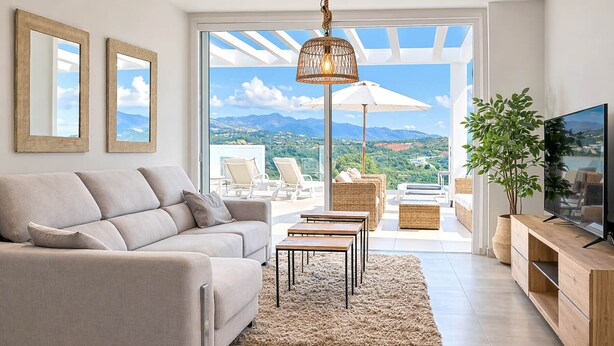 Un salon moderne avec un canapé, un téléviseur et une vue sur un paysage montagneux, visible à travers une grande fenêtre.