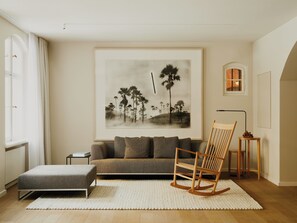 A living room with a sofa, a rocking chair, a rug, a framed picture on the wall, and a small table with a lamp.