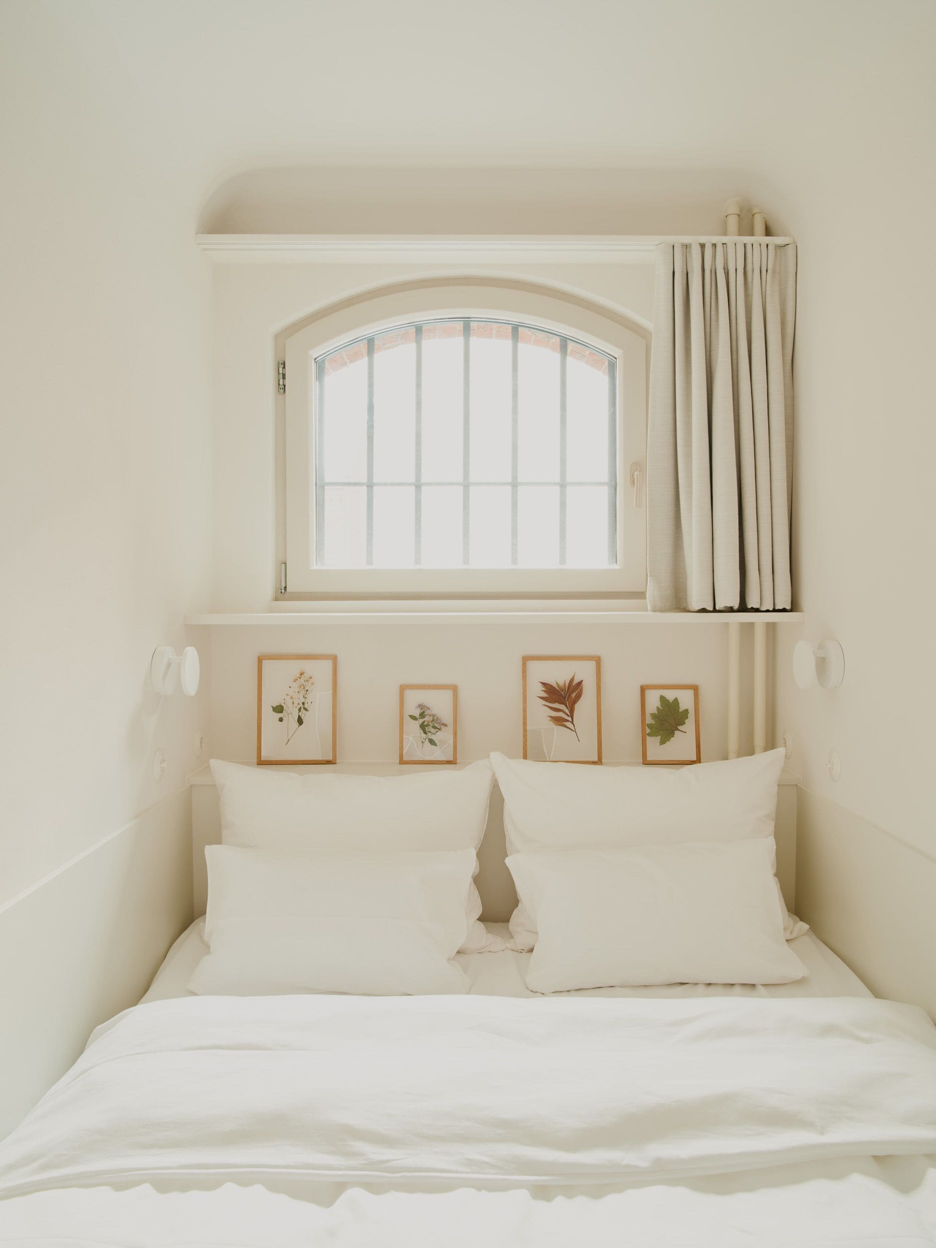 Una cama con ropa de cama blanca, una ventana con cortinas y tres láminas botánicas enmarcadas en la pared.