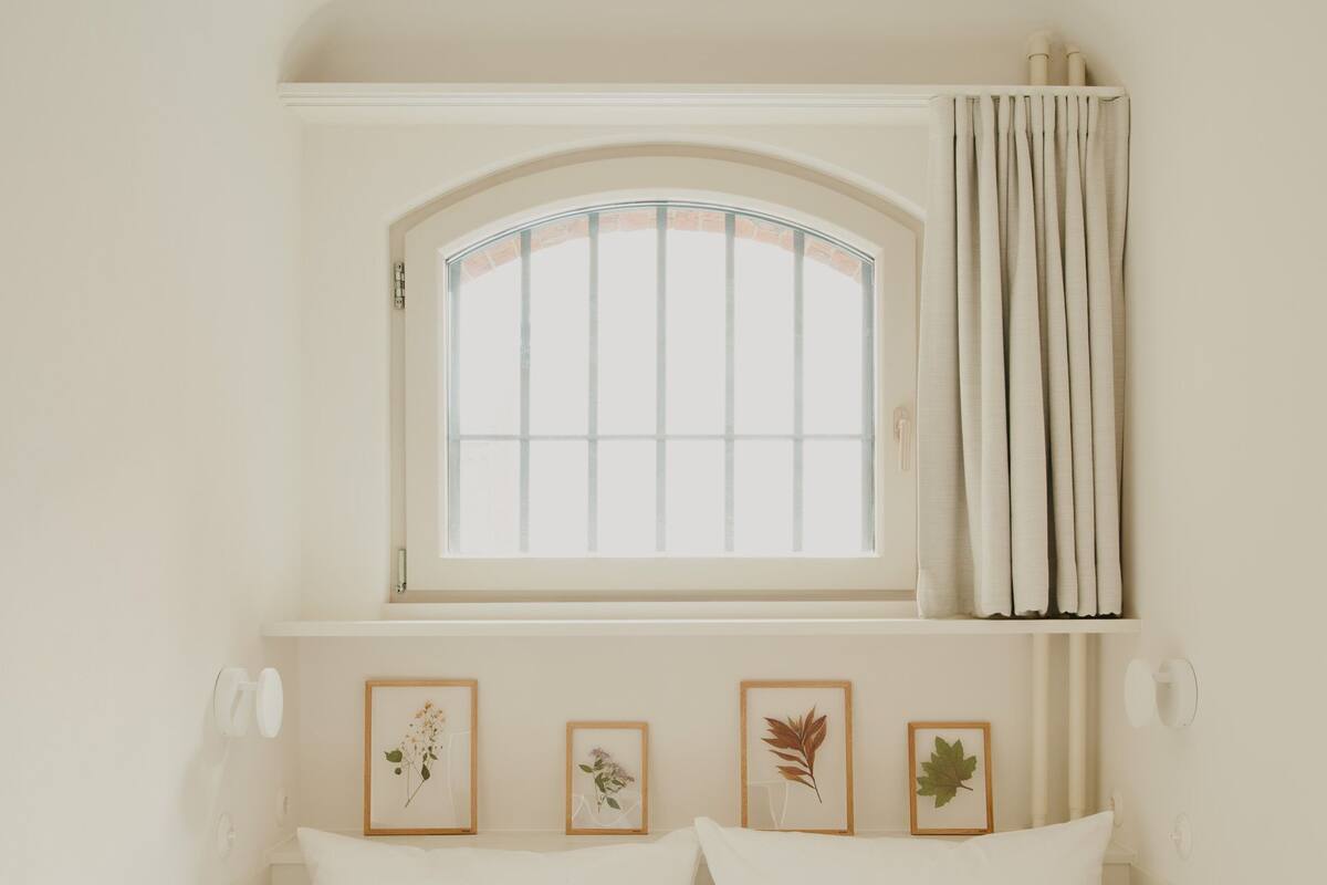 A bed with white bedding, a window with curtains, and three framed botanical prints on the wall.