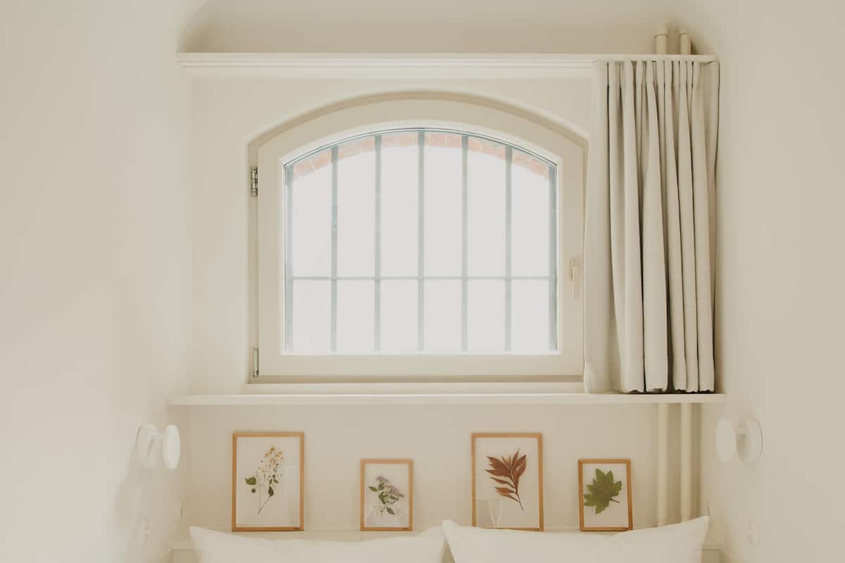 Una cama con ropa de cama blanca, una ventana con cortinas y tres láminas botánicas enmarcadas en la pared.