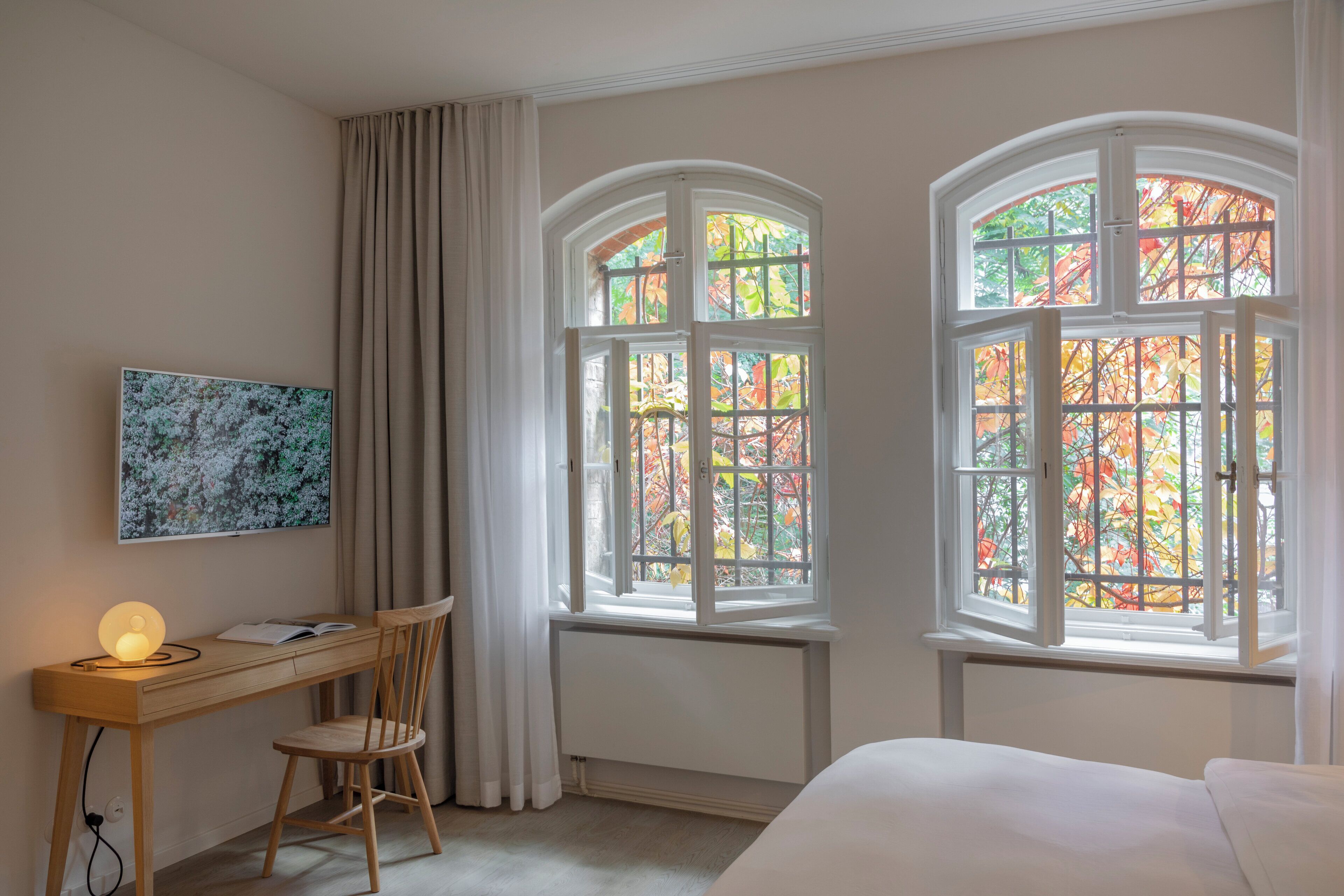 Un dormitorio con cama, un escritorio con silla, una ventana que ofrece vistas de hojas otoñales y un cuadro en la pared.
