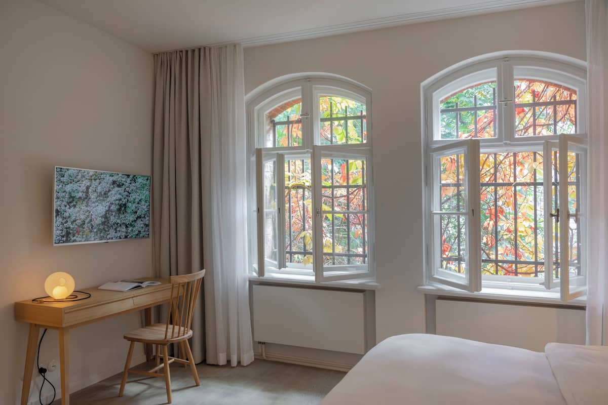 A bedroom with a bed, a desk with a chair, a window with a view of autumn leaves, and a painting on the wall.