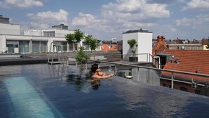 A rooftop swimming pool with a person relaxing, surrounded by modern buildings and clear skies.