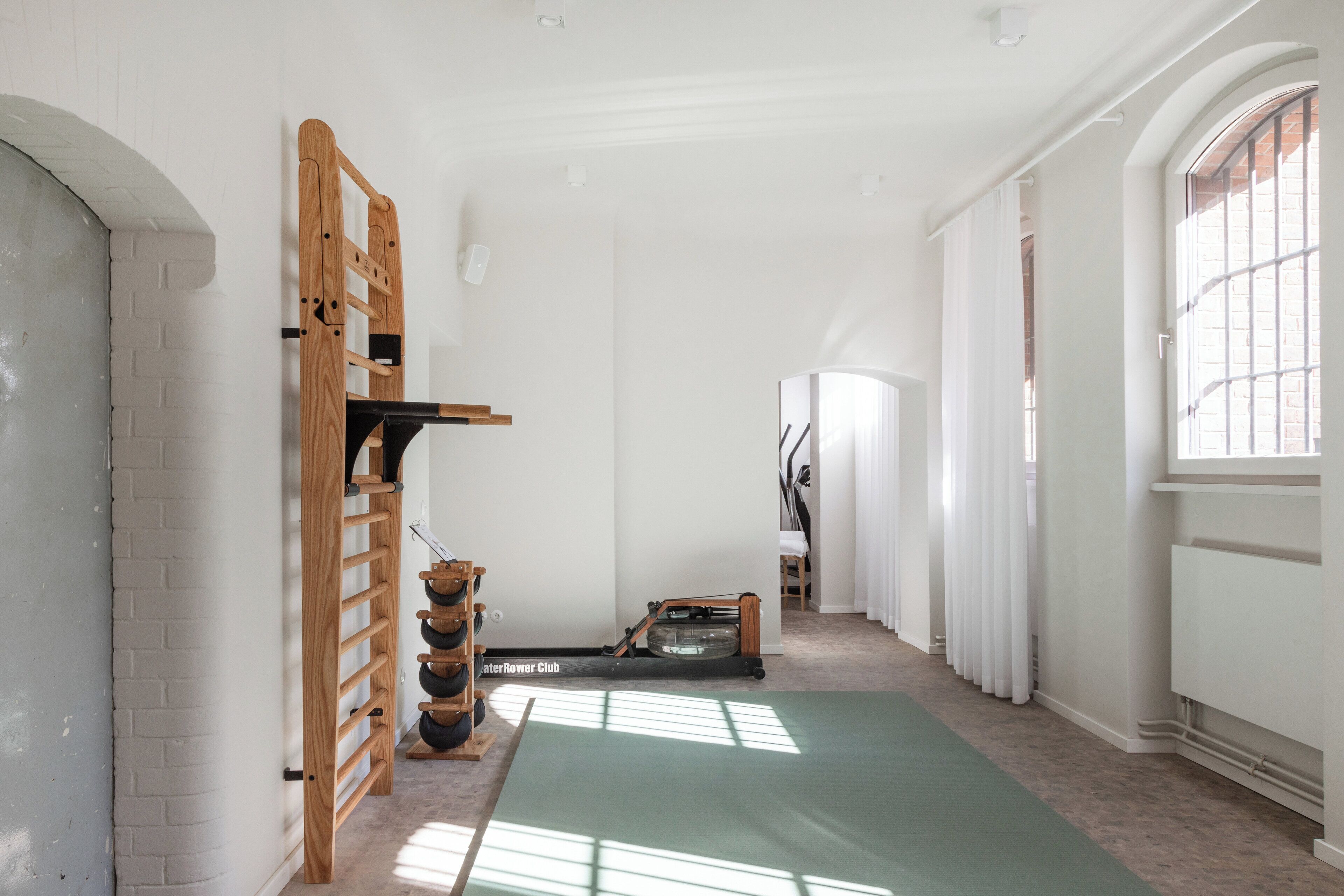 Een ruime kamer met een grote groene yogamat, een houten rek met zwarte gewichten, en een waterroeimachine.