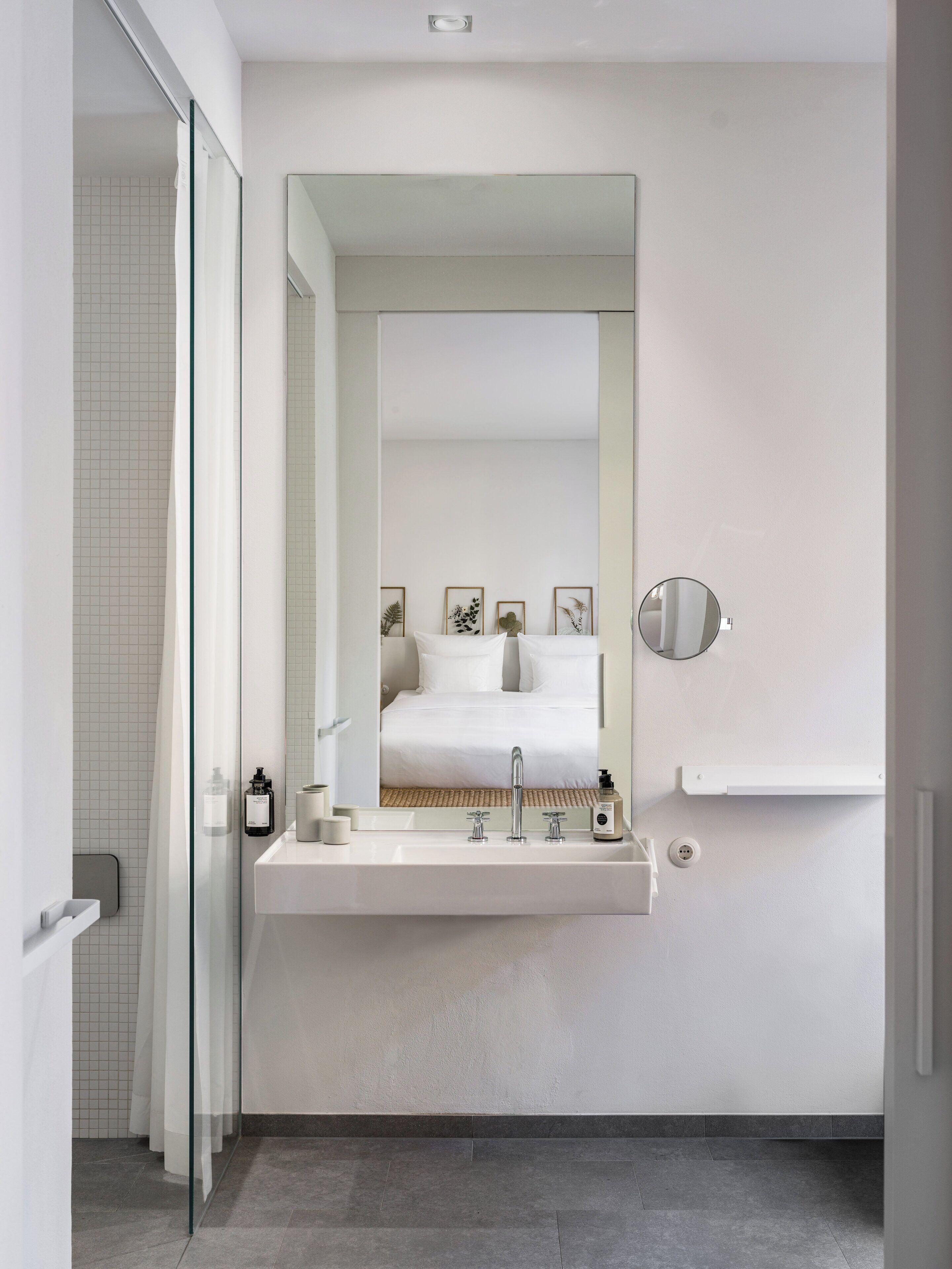 A modern bathroom with a glass shower enclosure, a built-in sink with a mirror, and a bed visible through a large mirror.