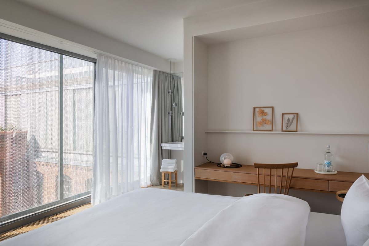 A hotel room with a large bed, a desk, and a chair. There is a window with curtains, a small stool, and a water bottle on the desk.
