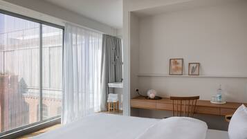 Habitación de hotel con una cama grande, un escritorio y una silla. Se observa una ventana con cortinas, un taburete pequeño y una botella de agua sobre el escritorio.