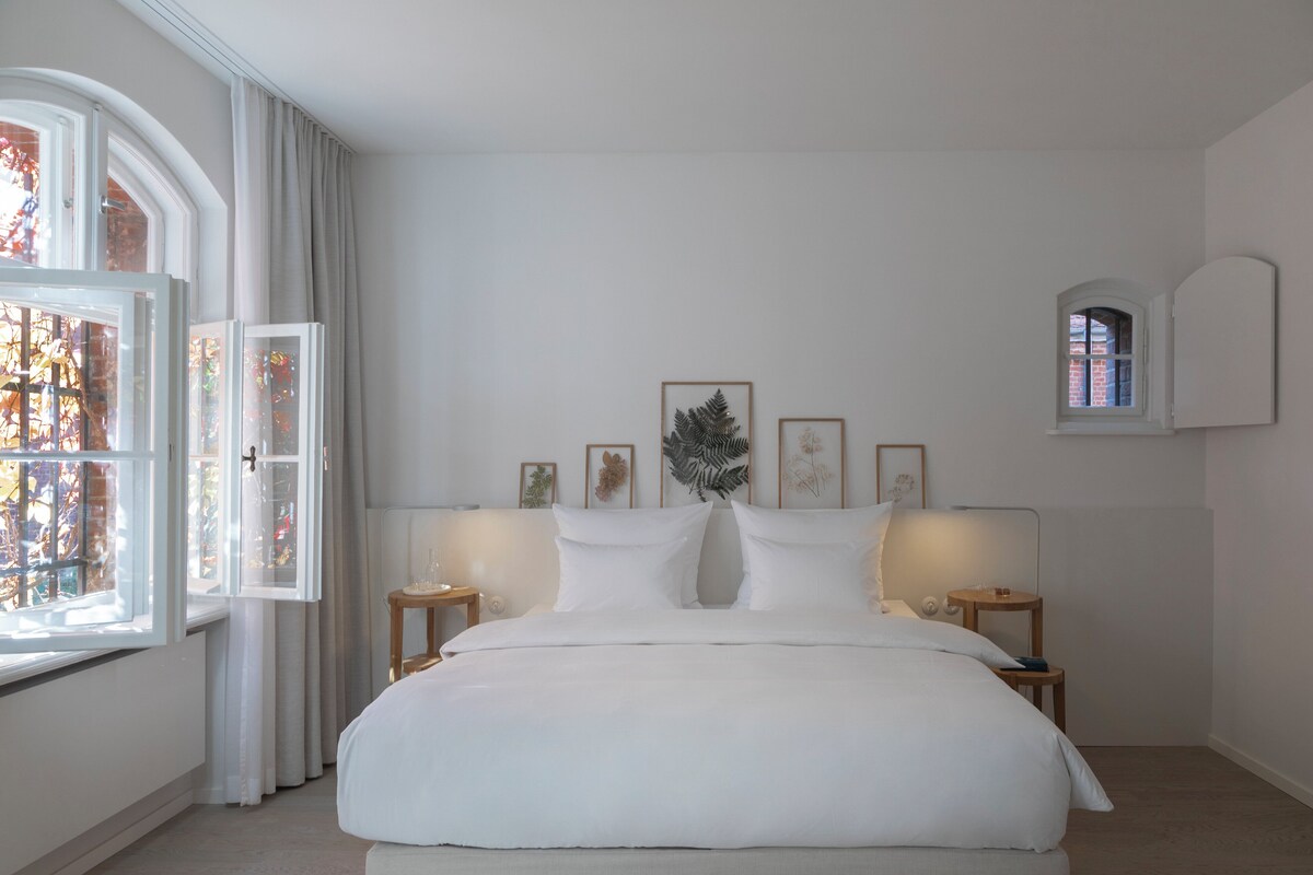 Un dormitorio con una cama grande, dos mesitas de noche, una ventana con cortinas y cuadros en la pared.