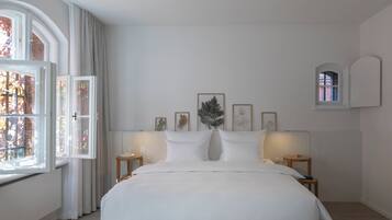 Une chambre à coucher avec un grand lit, deux tables de chevet, une fenêtre avec des rideaux et des œuvres d’art encadrées accrochées au mur.