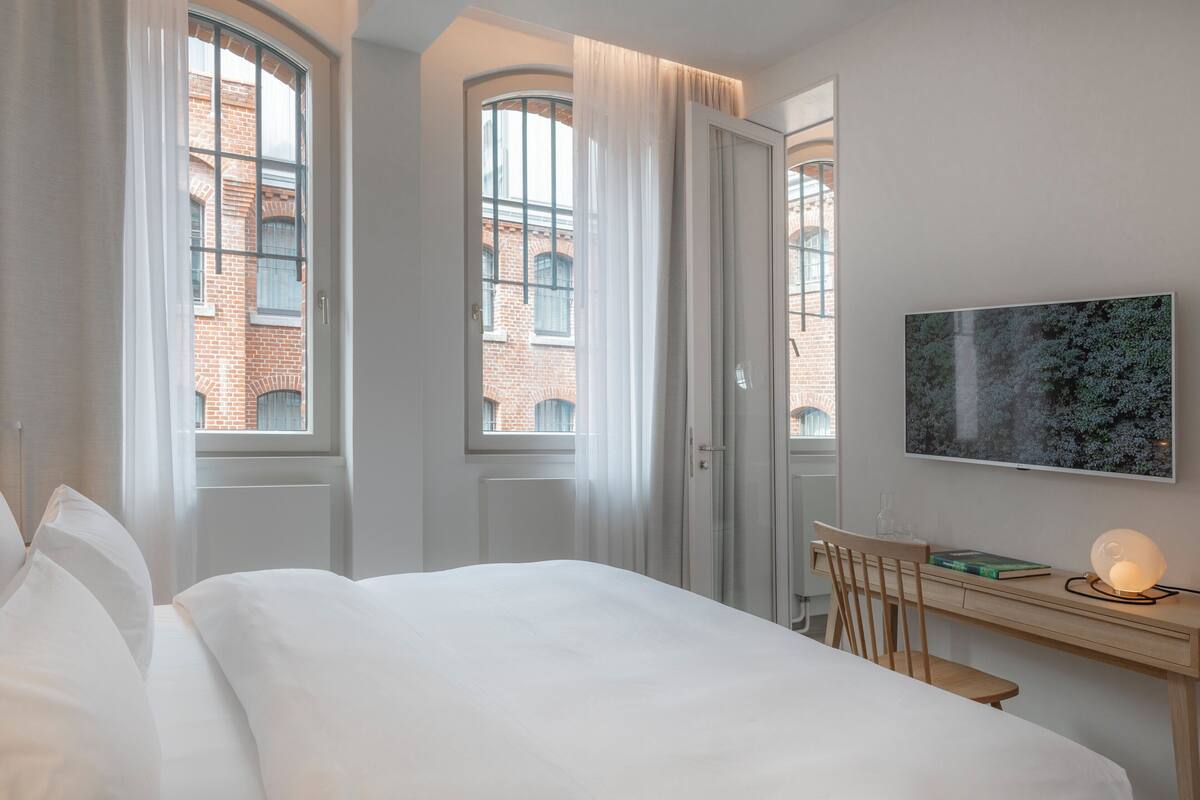 A hotel room with a bed, a desk, a chair, a TV, and large windows with curtains.