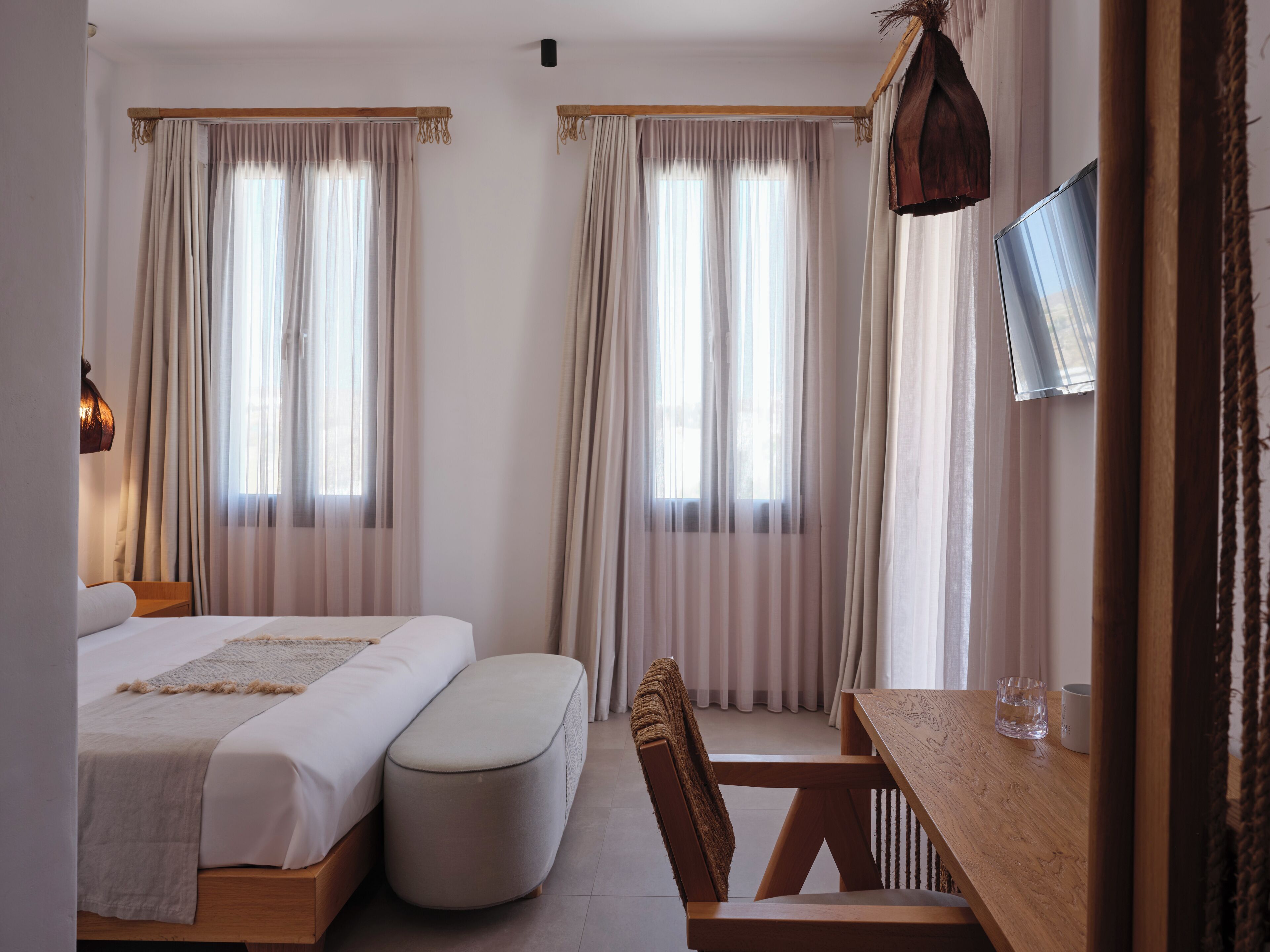 Une chambre à coucher avec un lit, un pouf, une table en bois, une chaise et un téléviseur fixé au mur.
