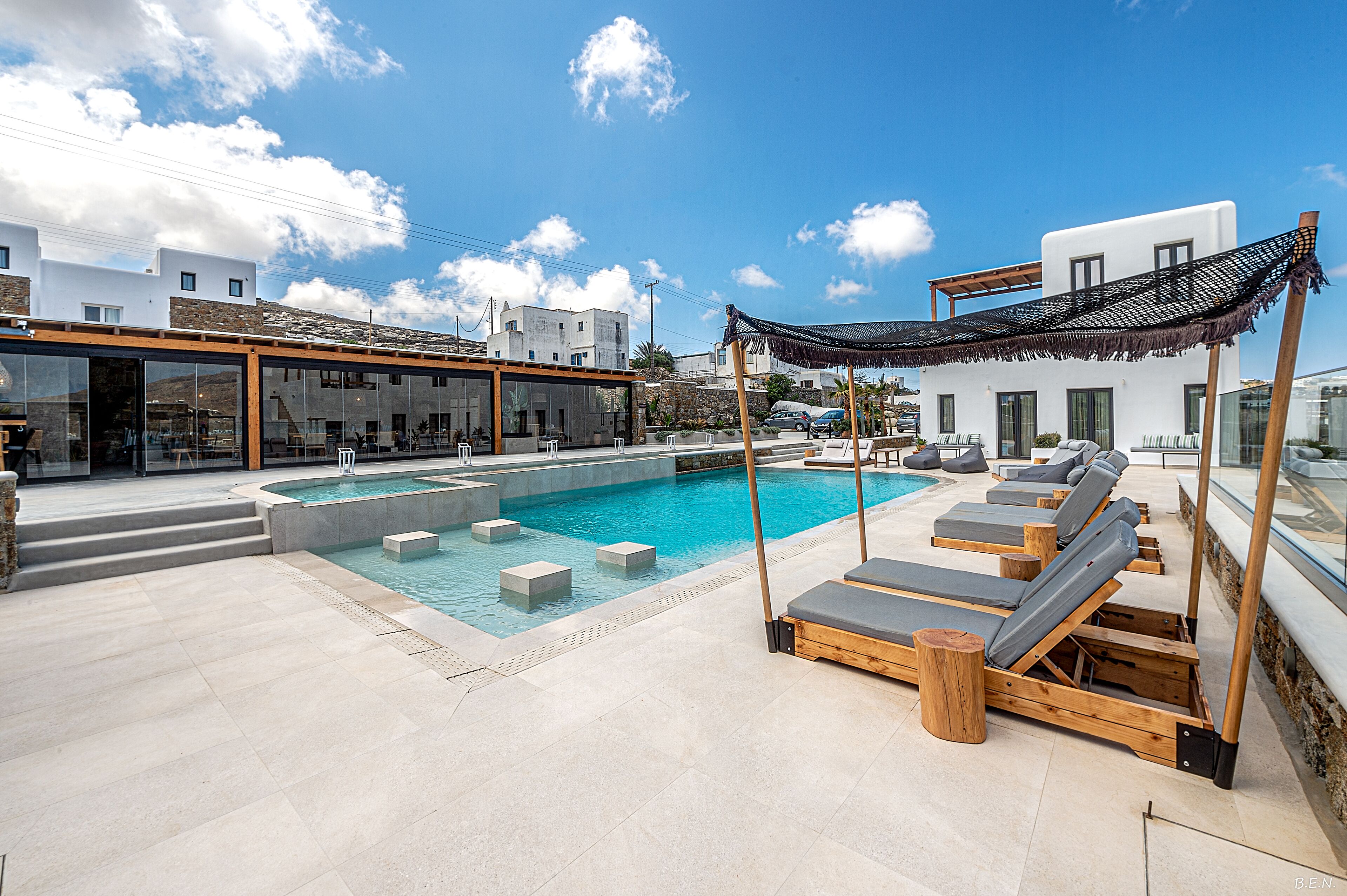 A rooftop pool area with a clear blue sky, a pool with floating steps, and wooden lounge chairs.