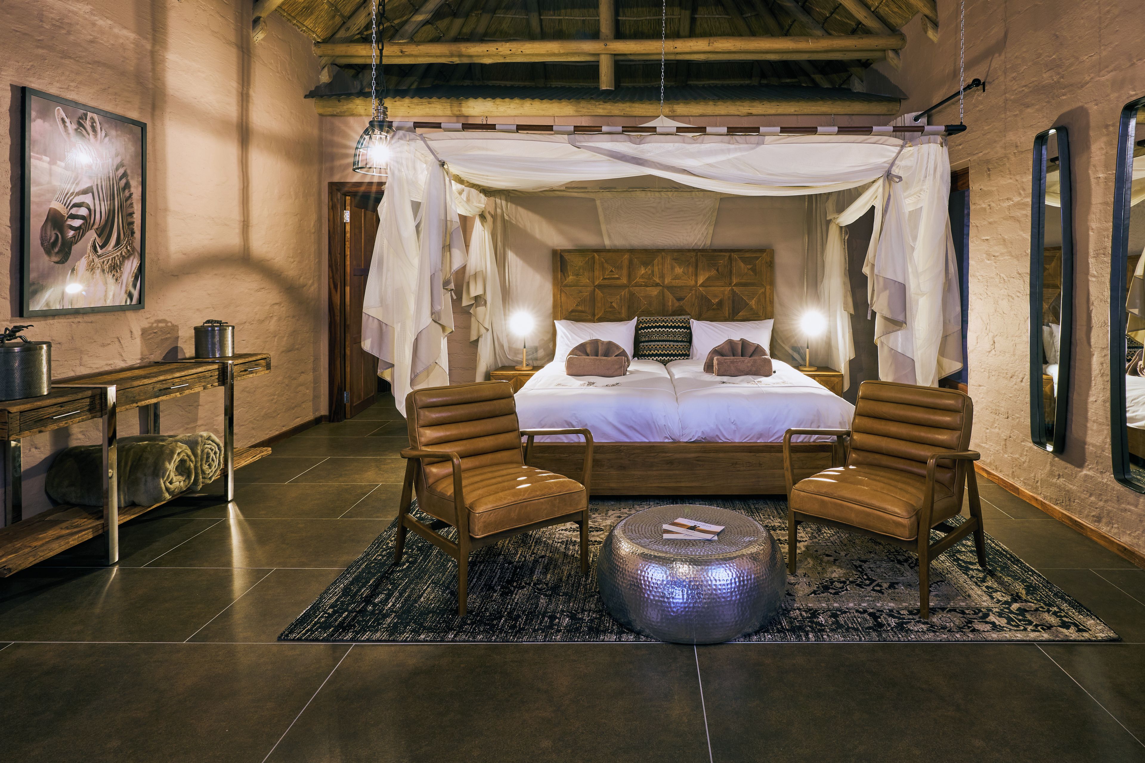Een ruime slaapkamer met een hemelbed, twee leren fauteuils, een rond pouf en een nachtkastje met een lamp.