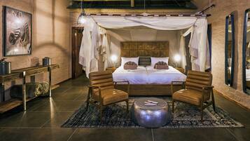 A spacious bedroom with a canopy bed, two leather chairs, a round ottoman, and a nightstand with a lamp.