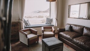 A living room with a brown leather sofa, two armchairs, a wooden coffee table, and a large window with a view of a desert landscape. - Desert Homestead Lodge l Ondili (Sesriem)