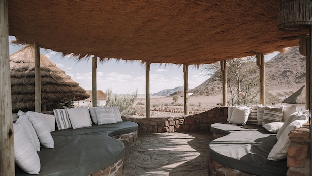 A covered outdoor seating area with stone walls and cushions, overlooking a desert landscape. - Desert Homestead Lodge l Ondili (Sesriem)