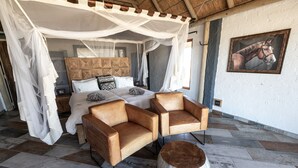 A bedroom with a four-poster canopy bed, two leather armchairs, a round coffee table, and a large painting on the wall. - Desert Homestead Lodge l Ondili (Sesriem)