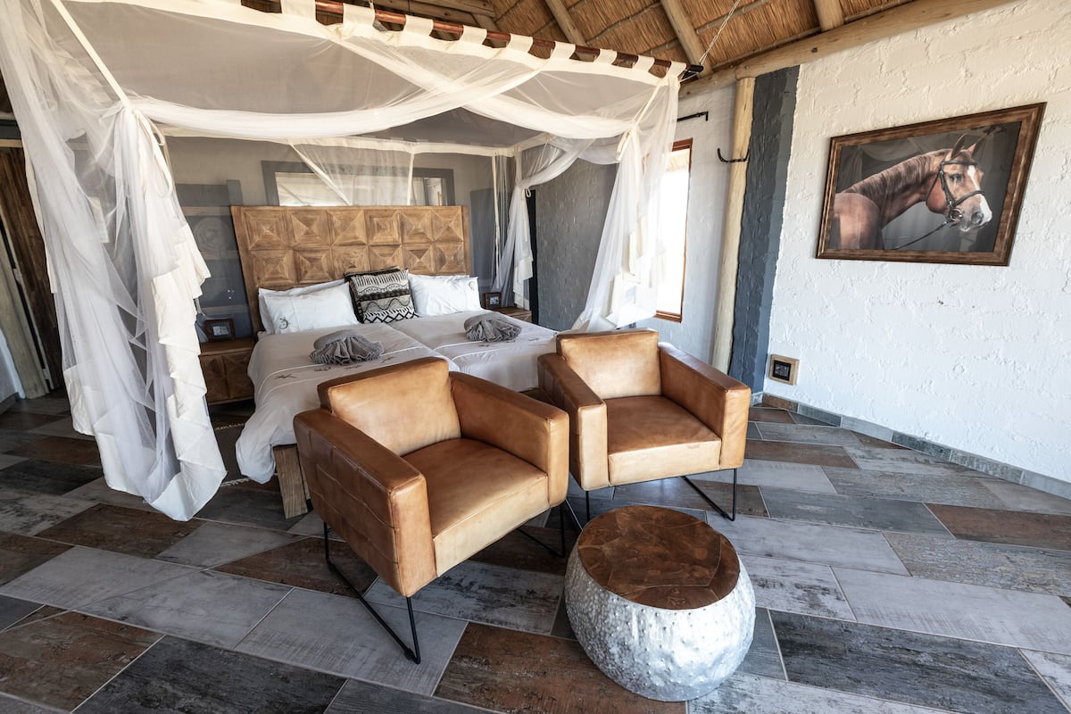 A bedroom with a four-poster canopy bed, two leather armchairs, a round coffee table, and a large painting on the wall.
