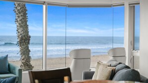 Ein Zimmer mit großem Fenster und Meerblick. Vor dem Fenster steht eine Palme, ein weißer Stuhl und ein Sofa mit Kissen.