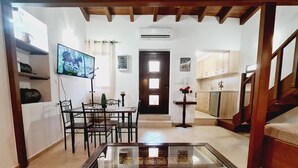 Un salon comprenant une table basse avec un plateau en verre, un téléviseur fixé au mur, un coin repas avec une table et des chaises, et un coin cuisine avec des placards et un plan de travail.