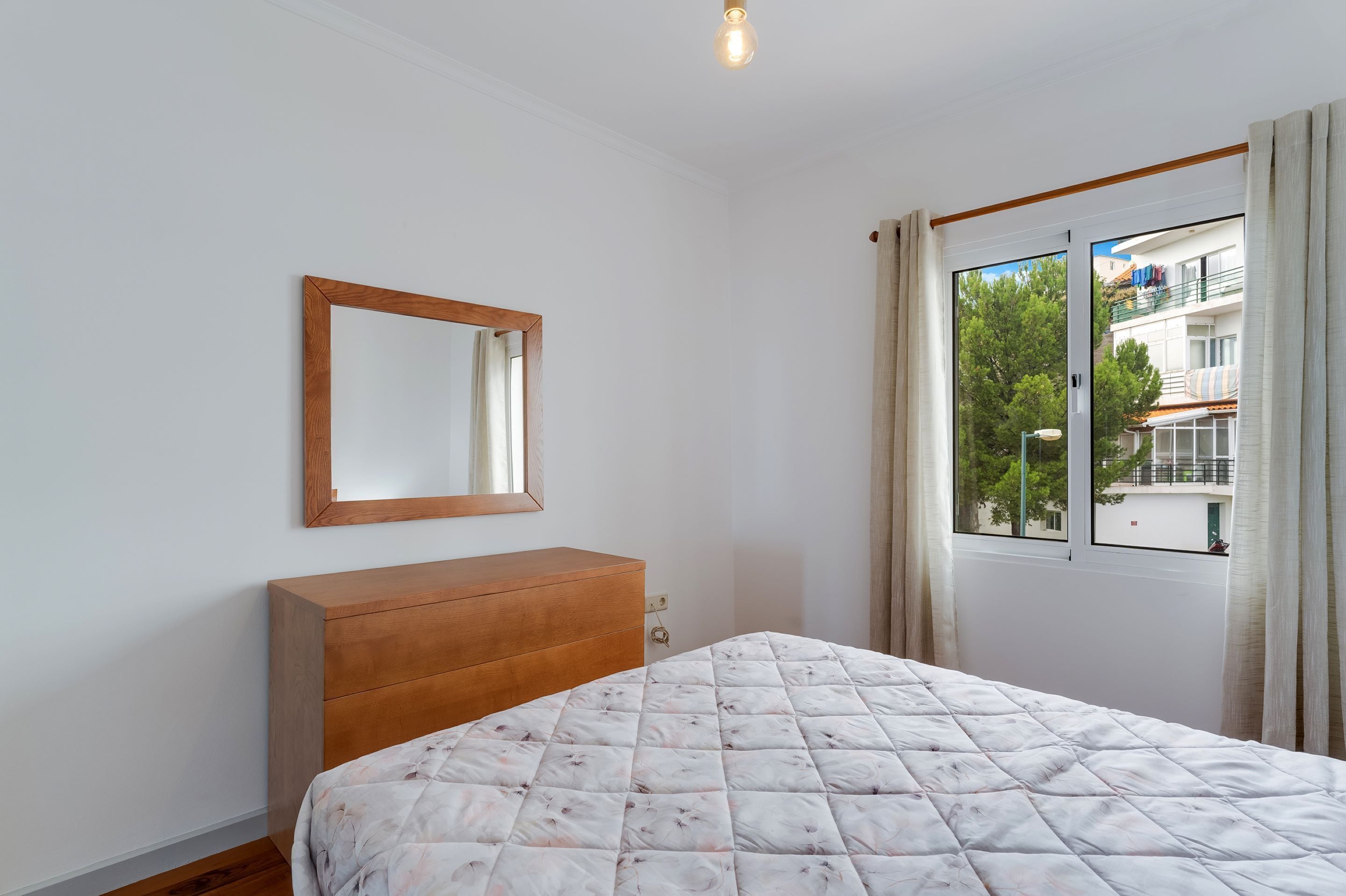 A bedroom with a wooden dresser, a bed with a floral quilt, a mirror, and a window with curtains.