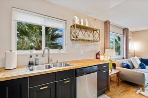 A modern kitchen with a wooden countertop, black cabinets, and stainless steel appliances. There is a window with a view of palm trees and a shelf with glassware above the sink.