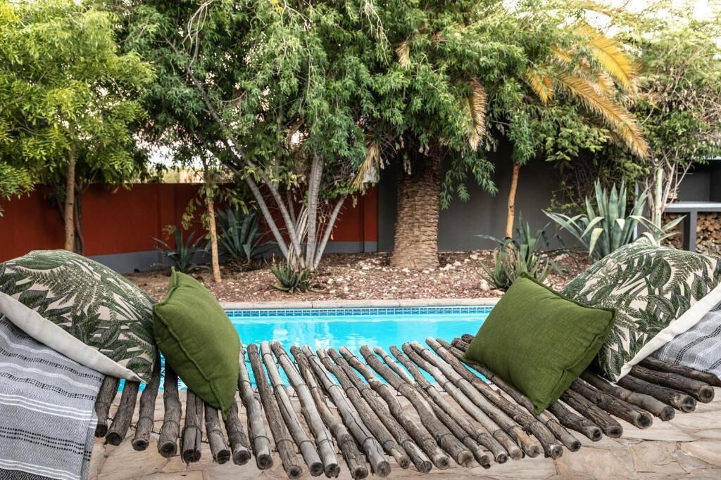 A poolside lounge area with a bamboo mat and pillows, surrounded by trees and a red wall.