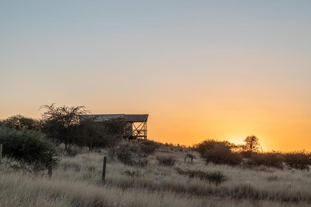 A rustic wooden structure with a metal framework, surrounded by dry grass and sparse vegetation, under a clear sky.