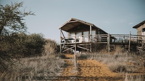 Eine rustikale Holzhütte mit einem Balkon, umgeben von trockenem Gras und Büschen.