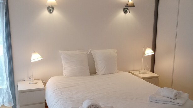 Un lit bien fait, avec du linge de lit blanc et deux lampes de chevet, un plancher en bois et une armoire blanche, dans une chambre d'hôtel.