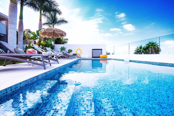 A swimming pool with lounge chairs and umbrellas, surrounded by palm trees and a clear sky.