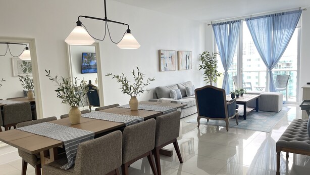 A modern living room with a dining area, featuring a sofa, a coffee table, and a dining table with chairs. There are potted plants and artwork on the walls.