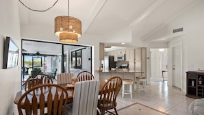 A dining area with a table and chairs, a kitchen in the background, and a large window.