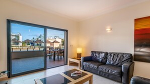 A modern living room with a glass sliding door leading to an outdoor terrace, a leather sofa, a wooden coffee table, and a cityscape view.