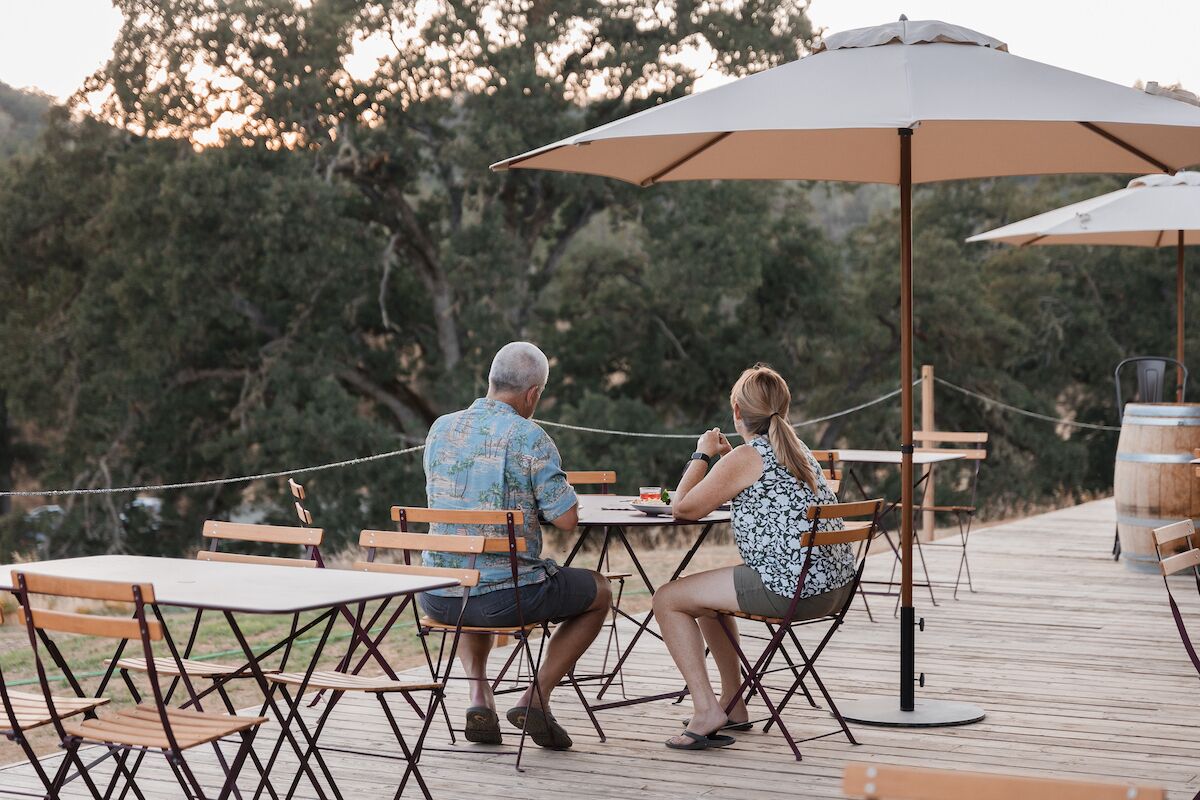 A couple dining at an outdoor table with a large umbrella, surrounded by wooden chairs and tables on a wooden deck.