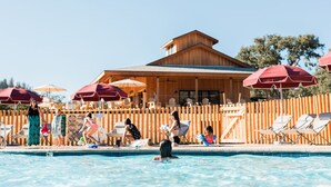 A pool area with wooden deck chairs and umbrellas, a wooden building with a sloped roof, and a clear blue sky. - Huttopia Wine Country-Northern California (Lower Lake)