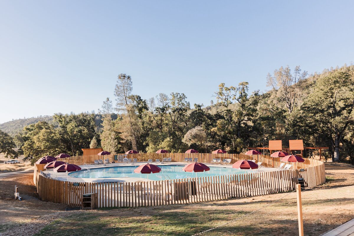 A circular pool with wooden fencing and red umbrellas, surrounded by trees and hills.