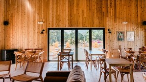 A wooden interior with tables and chairs arranged for dining, large windows with a view of the outdoors, and a ceiling fan. - Huttopia Wine Country-Northern California (Lower Lake)