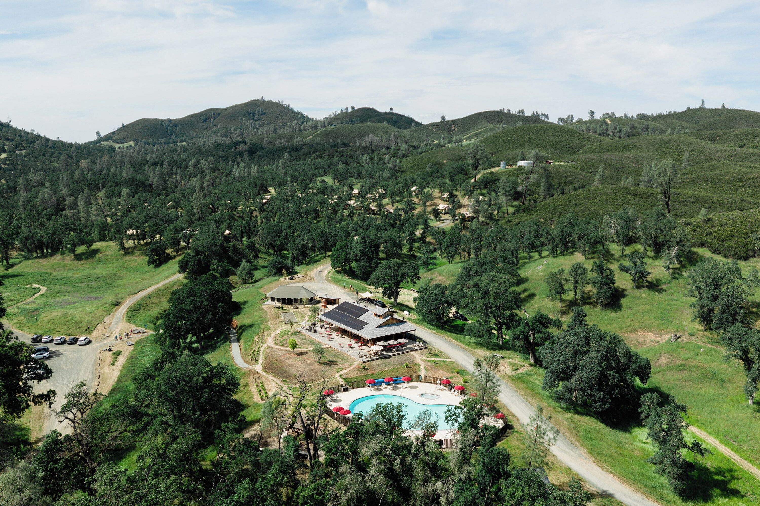 An aerial view of a resort with a swimming pool, guest rooms, and a dining area surrounded by hills and trees.