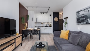 Un salon moderne avec un canapé, une table basse et un téléviseur. On y trouve également un coin repas avec des chaises et une table. La cuisine est visible au fond.